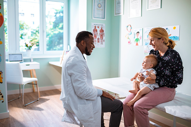 A baby sits on their mother's lap in a pediatrician's office as they both look up at a happy doctor