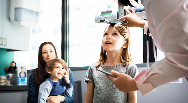 A pediatrician measures a young girl's height as the girl's mother and younger brother watch
