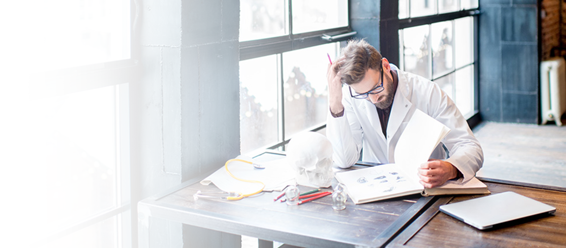 Doctor studies a textbook at a desk near many windows