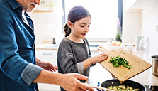 Young girl cooking vegetables with her parent overseeing