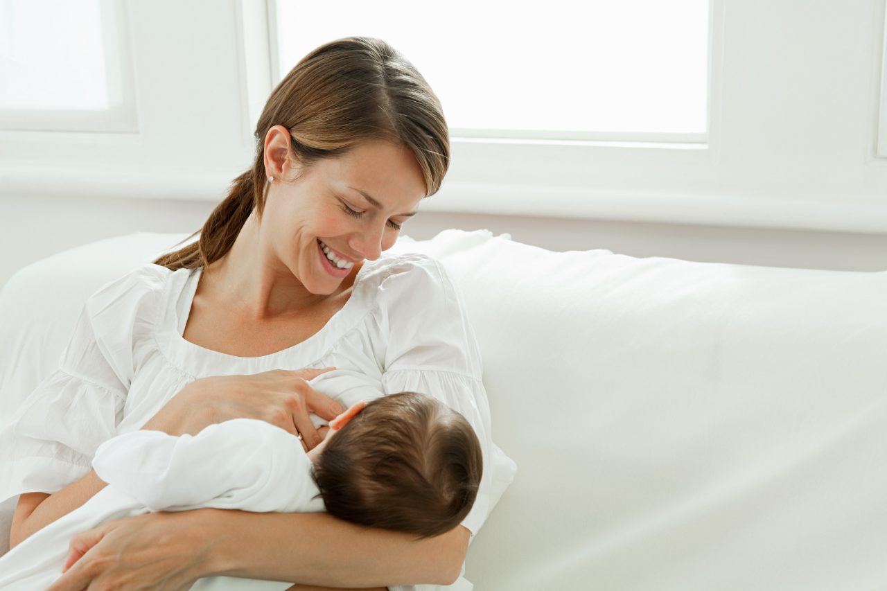 Mother breastfeeding baby while sitting on white couch Breastfeeding Education: Tips & Techniques for Getting Started